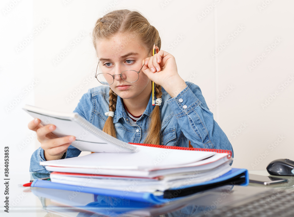 Naklejka premium Concentrated young girl doing her homework with laptop and notebook at kitchen table