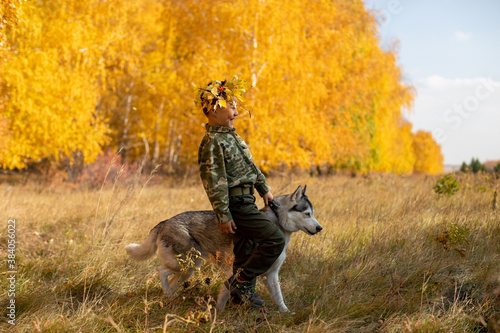 Smiling boy wearing khaki and an autumnal foliage crown walking in the countryside with dog on the background of the autumn birch forest.