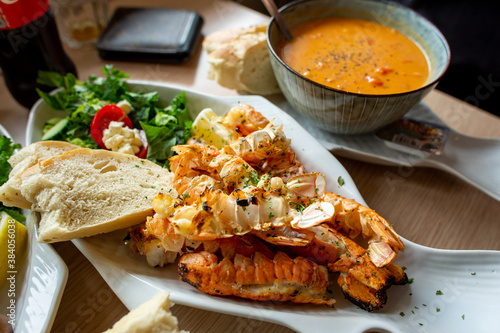 Langoustine is grilled on a white fish-shaped platter. Set beautiful breads and vegetables on the dining table. There is a bowl of soup on the side. At a restaurant in the southern city of Iceland