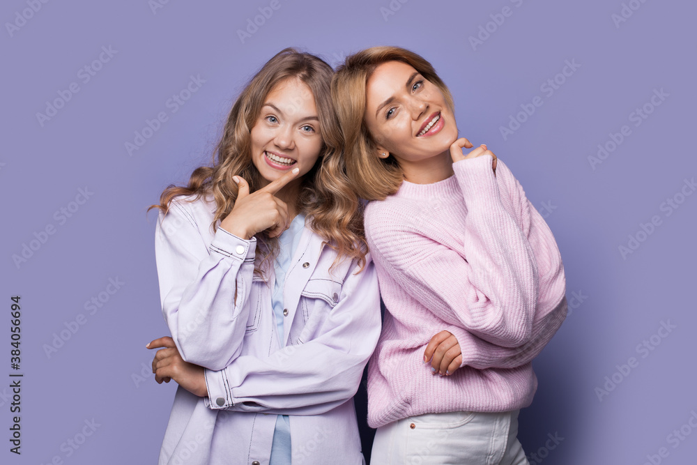Blonde sisters smiling at camera in casual clothes touching their chin on a violet studio wall Blonde sisters smiling at camera in casual clothes touching their chin on a violet studio wall