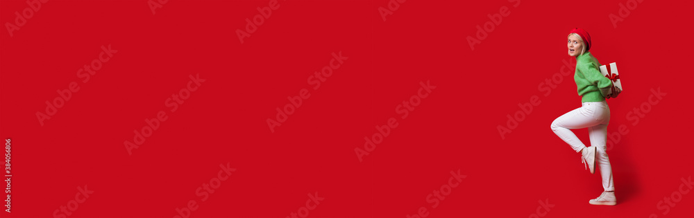 Blonde caucasian woman is carrying a present box on her back wearing a hat and looking at camera on a red wall with free space Blonde caucasian woman is carrying a present box on her back wearing a hat and looking at camera on a red wall with free space