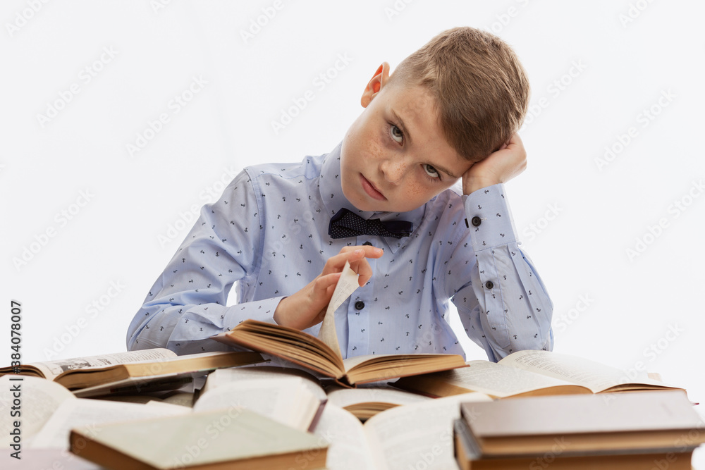 A sad tired schoolboy in a blue shirt with a bow tie sits over textbooks. Back to school. White background. A sad tired schoolboy in a blue shirt with a bow tie sits over textbooks. Back to school. White background.