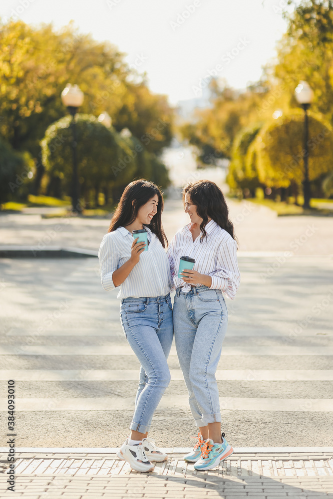 Two people women friends in jeans and shirts are standing near the door drinking coffee and chatting outdoors