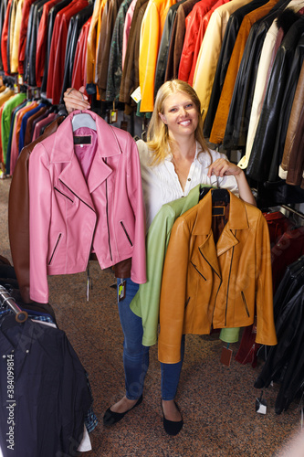 Attractive young blonde woman choosing leather jacket on racks in clothes store