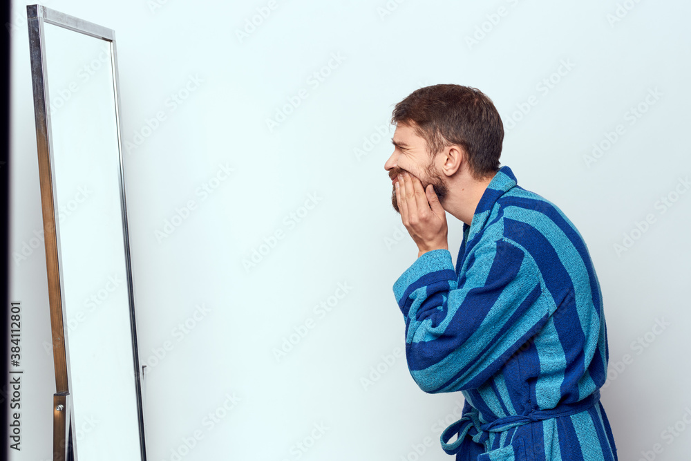 a man in a blue robe examines himself in a mirror in a bright room cropped view a man in a blue robe examines himself in a mirror in a bright room cropped view