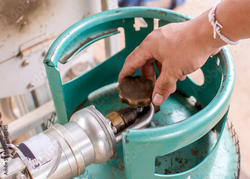 The silver dispenser is connected to the gas cylinder valve and the male operator's hand turns to open the gas cylinder refill valve.