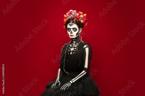 Scary. Young girl like Santa Muerte Saint death or Sugar skull with bright make-up. Portrait isolated on red studio background with copyspace. Celebrating Halloween or Day of the dead.