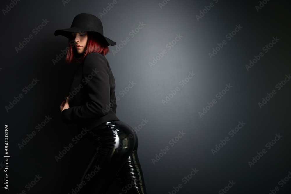 Beautiful fashion woman with perfect body. Portrait of beautiful girl wearing black blazer, latex tight pants and hat posing in studio over deep gray background Beautiful fashion woman with perfect body. Portrait of beautiful girl wearing black blazer, latex tight pants and hat posing in studio over deep gray background