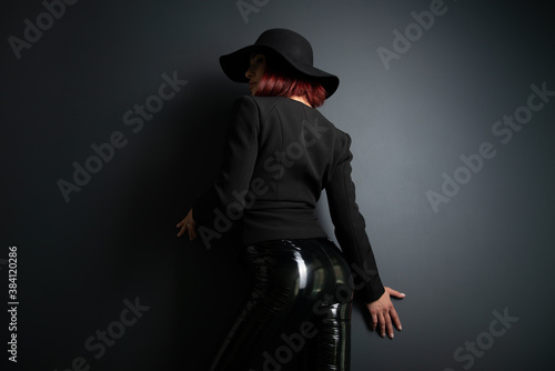 Beautiful fashion woman with perfect body, back view. Portrait of beautiful girl wearing black blazer, latex tight pants and hat posing in studio over deep gray background