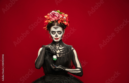 Gambling. Young girl like Santa Muerte Saint death or Sugar skull with bright make-up. Portrait isolated on red studio background with copyspace. Celebrating Halloween or Day of the dead.