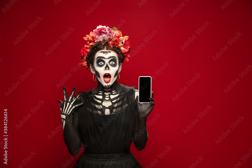 Holding smartphone. Young girl like Santa Muerte Saint death or Sugar skull with bright make-up. Portrait isolated on red studio background with copyspace. Celebrating Halloween or Day of the dead.