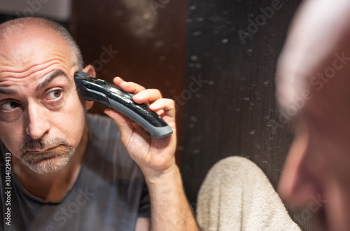 A middle-aged Caucasian man cuts his hair in the bathroom. While placing the clipper on your head check with your eyes in the mirror.