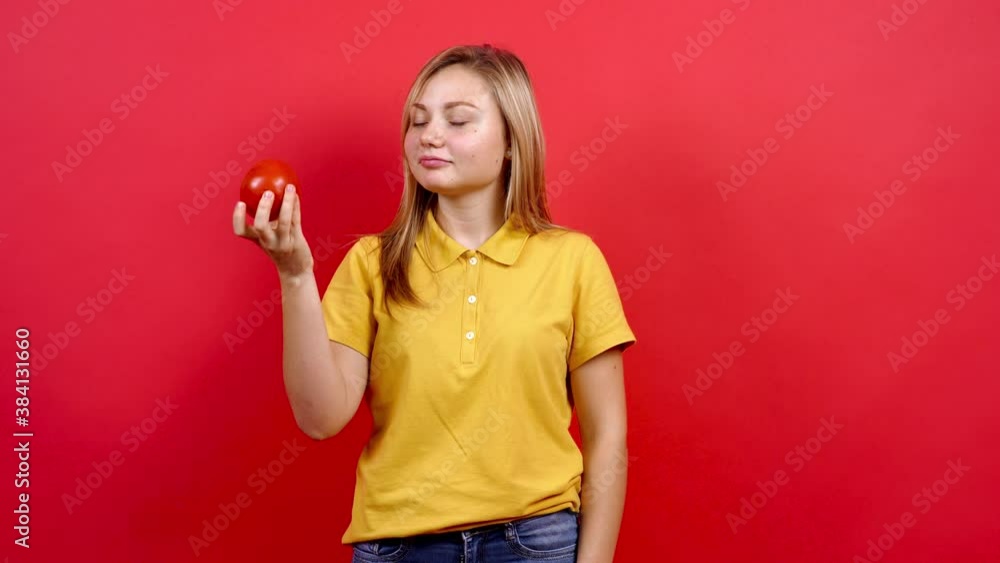 Cute and slightly fat girl in a yellow T-shirt who holds a fresh tomato in her hand, recommends the consumption of fruits and vegetables, healthy nutrition. indoor studio shot isolated on red Cute and slightly fat girl in a yellow T-shirt who holds a fresh tomato in her hand, recommends the consumption of fruits and vegetables, healthy nutrition. indoor studio shot isolated on red