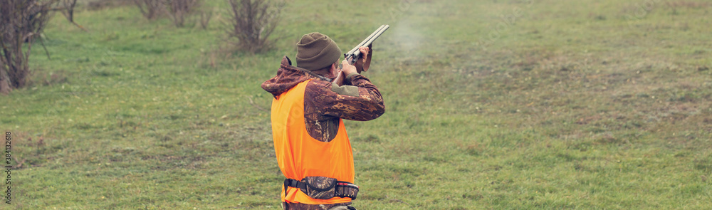 Fototapeta premium A man with a gun in his hands and an orange vest on a pheasant hunt in a wooded area in cloudy weather. Hunter with dogs in search of game.