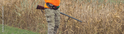 A man with a gun in his hands and an orange vest on a pheasant hunt in a wooded area in cloudy weather. Hunter with dogs in search of game.