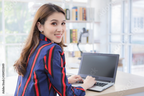 Asian confident woman is resting her hand on the keyboard of laptop and turn back her face with smiling in a working room at home.