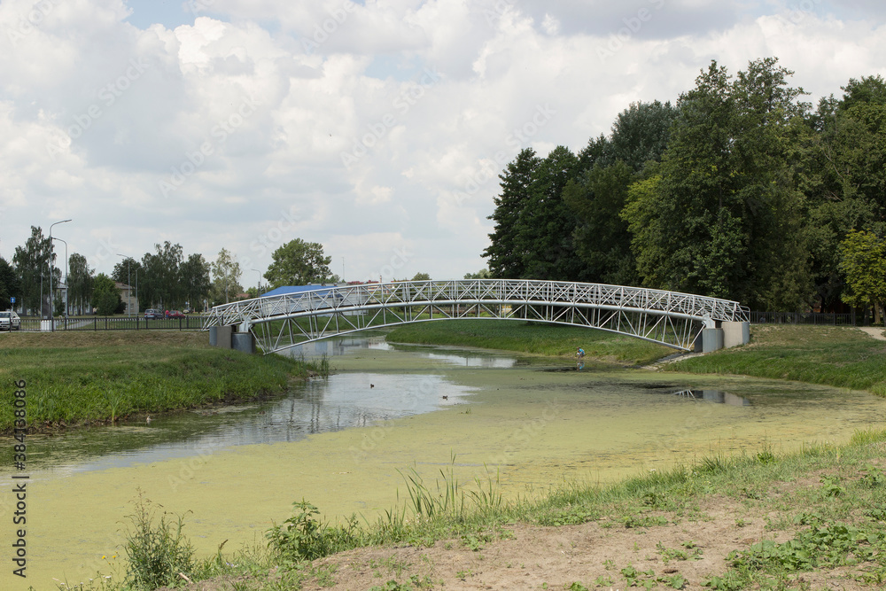 A modern metal bridge over a green city river with blooming algae. Unkempt river in the center of the park in summer. A modern metal bridge over a green city river with blooming algae. Unkempt river in the center of the park in summer.