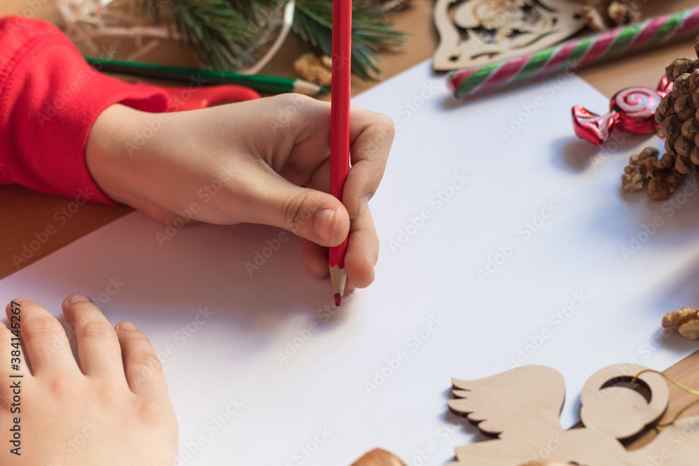 Children's hand writes with a pencil. Child's hands, the sheet of paper, pencils and Christmas decorations on a wooden surface. Concept of New year and Christmas. Side view