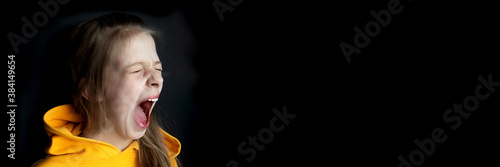 Teenager girl in an orange sweatshirt with bright emotions on her face on a black background. Girl screaming. Banner. Selective focus. Side view