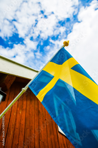 Swedish flag blowing in the wind. Red house in background. Blue cloudy sky. Selective focus on subject.
