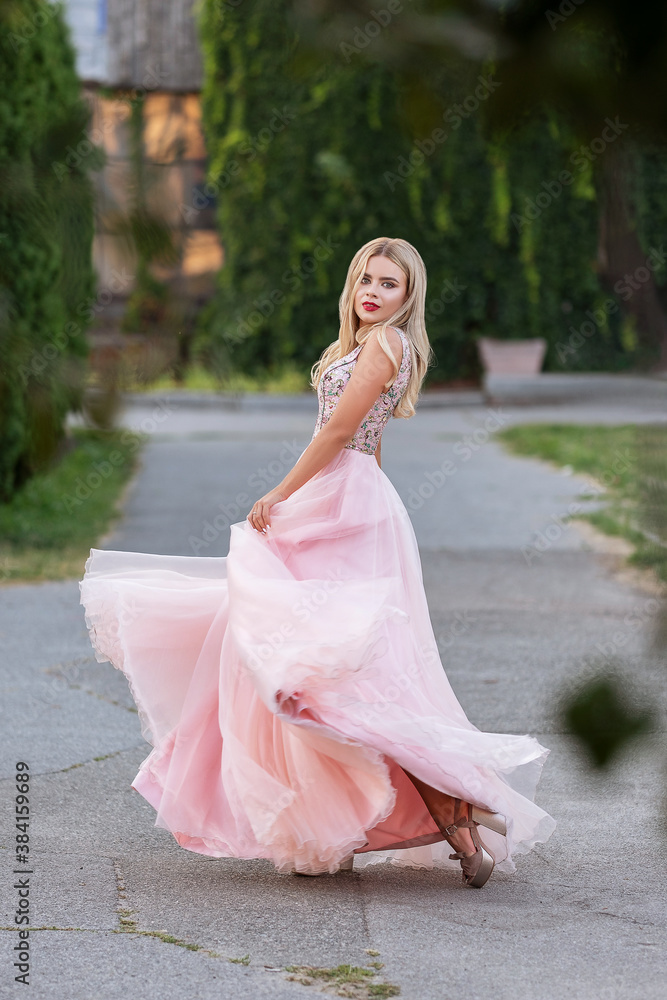 Blonde woman with make up in evening dress at park. The skirt of her dress fly on a wind. Sensual fashion portrait with a sunset light