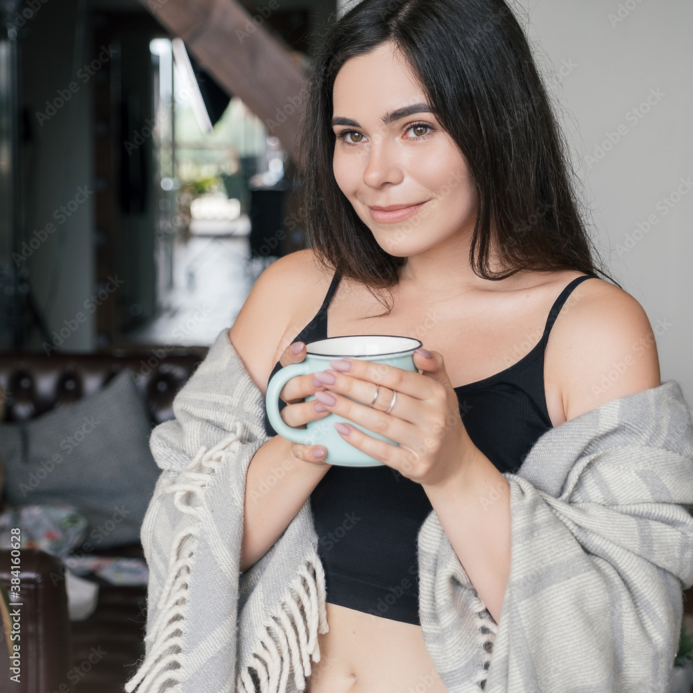 Smiling brunette woman at home covered in plaid drinking morning coffee near window. Smiling brunette woman at home covered in plaid drinking morning coffee near window.