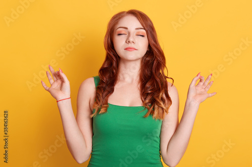 Wallpaper Mural Red haired peaceful Caucasian woman in green t shirt meditates indoor against yellow wall, keeps hands in mudra gesture, has eyes closed, relaxes after long hours working, holds fingers in yoga sign Torontodigital.ca