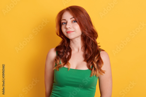 Portrait of satisfied red haired female model with curls, gentle smile, dressed in casual green sleeveless t shirt, looks straightly at camera, pose over yellow wall.