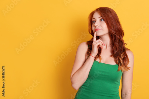 Beautiful red haired female with thoughtful expression, keeps finger on lips, looks aside with bewilderment, poses against yellow background, wearing green t shirt.
