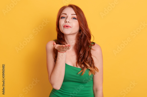 Adorable young woman with long red hair blows kiss, demonstrates her good feelings, says goodbye on distance, isolated over yellow background, attractive pretty female model makes air kiss.