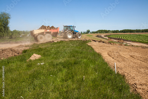 Tractor with tipper trailer loaded with clay drives down a dike on a dusty track; in the background a green polder and a reenforced dike with a new layer of clay. 