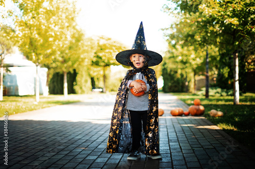 surprised child boy stands in the street dressed as a wizard in a hat and cloak holding a pumpkin in his hands. halloween. pumpkin
