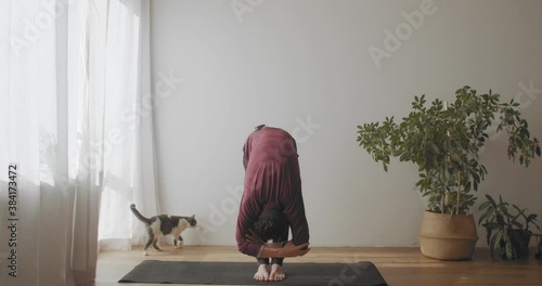 Fit man standing in uttanasana pose on mat indoors natural light slow motion. Yogi master demonstrating flexible body practicing yoga in sunny room copy text space. Active healthy lifestyle concept photo