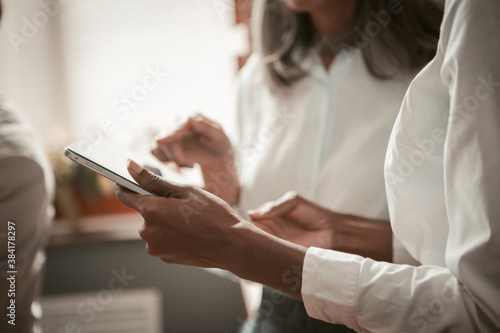 Businesswomen using digital tablet in office. Close up shot of female hands. African American and Asian women wearing white shirts backlit by sunlight. High quality photo. © Svyatoslav Lypynskyy