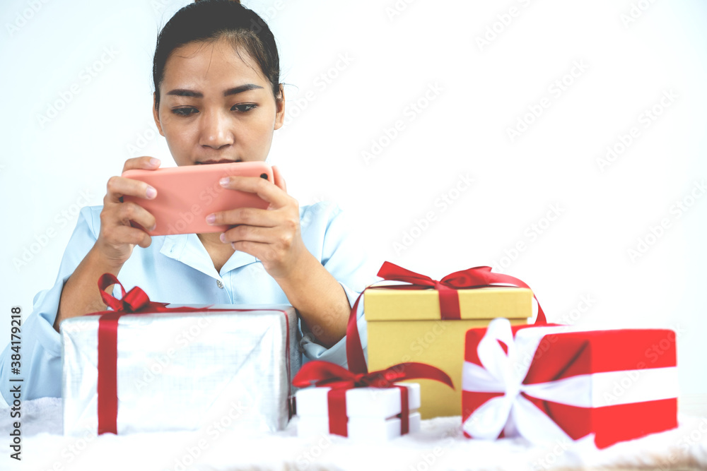 Woman in a blue shirt uses a smartphone to check the address for sent gift boxes tied with a red ribbon present for the festival of giving special holidays like Christmas, Valentine's Day Woman in a blue shirt uses a smartphone to check the address for sent gift boxes tied with a red ribbon present for the festival of giving special holidays like Christmas, Valentine's Day