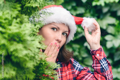 Surprised face woman in a santa claus red hat looks away through green fir tree branches in the forest. Christmas coming  sale concept.