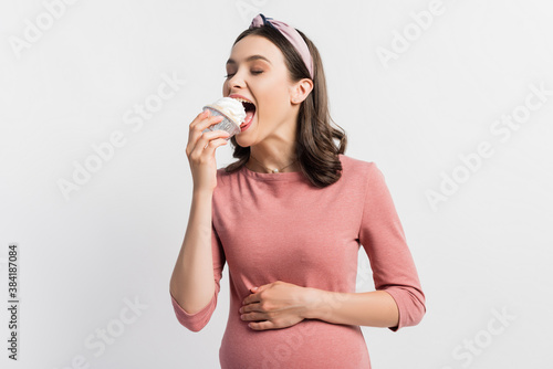 joyful pregnant woman with closed eyes eating cupcake isolated on white