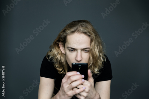Young man with long blonde hair taking pictures of something with a phone with a strained expression on his face on a black background