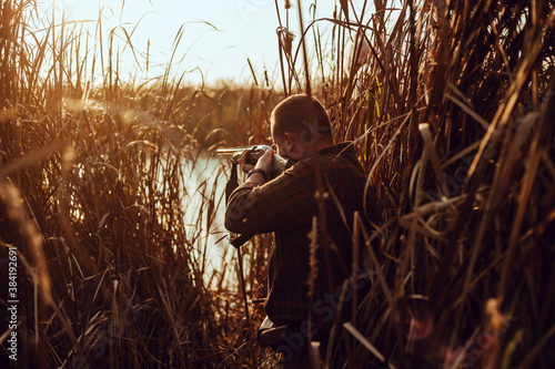 Young hunter man with a shotgun hiding in the reeds near the pond, at the duck hunt - photo with selective focus