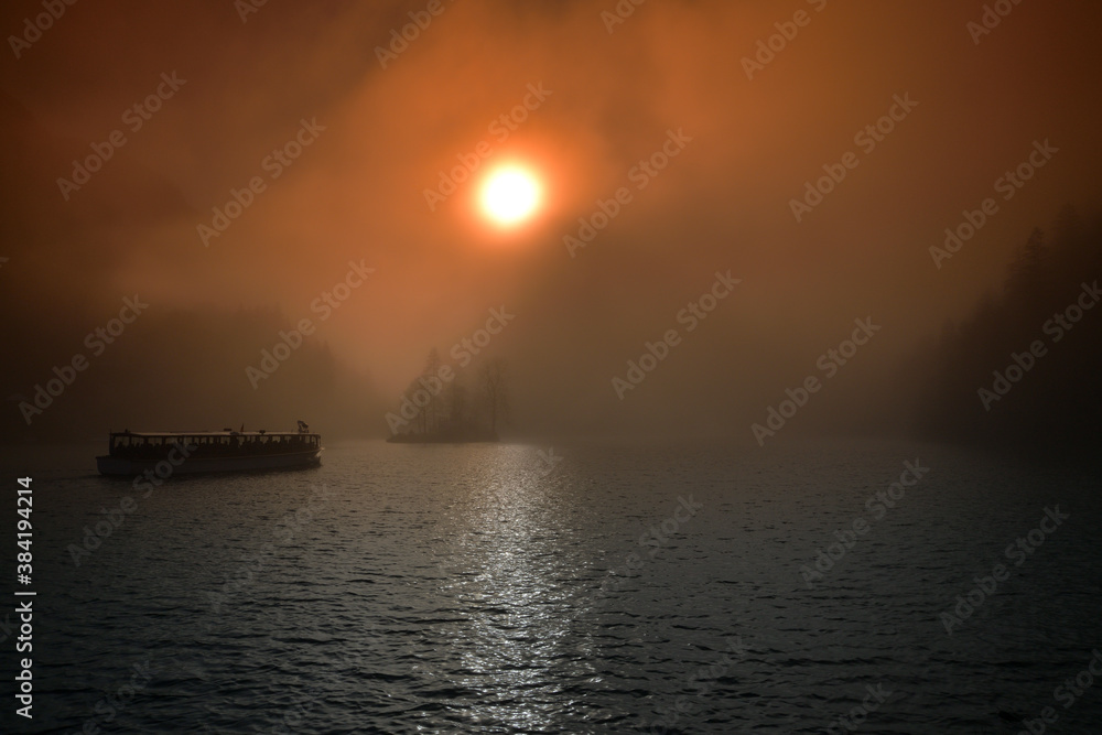 Sonnenaufgang über dem nebeligen Königssee mit einem fahrenden Schiff