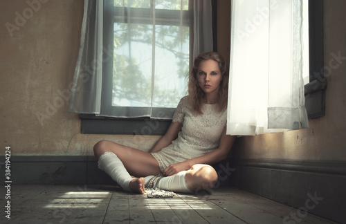 Beautiful young blonde woman wearing a white lace outfit and leg warmers, holding a pearl necklace while sitting alone in the corner of an empty old room, white curtains hanging from the windows photo