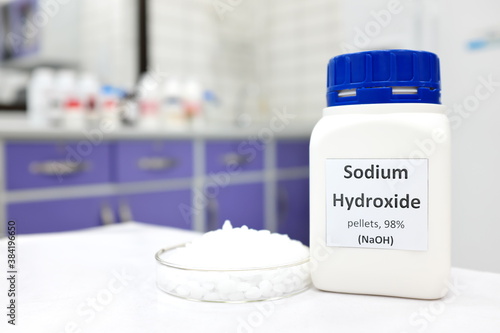 Selective focus of a bottle of pure sodium hydroxide or NaOH chemical compound beside a petri dish with white solid pellets. Chemistry research laboratory background with copy space. photo