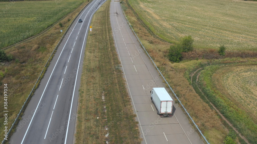 Aerial view of truck with cargo trailer driving on straight road and transporting goods. Camera following to delivery lorry moving across country highway. Flying over traffic in rural