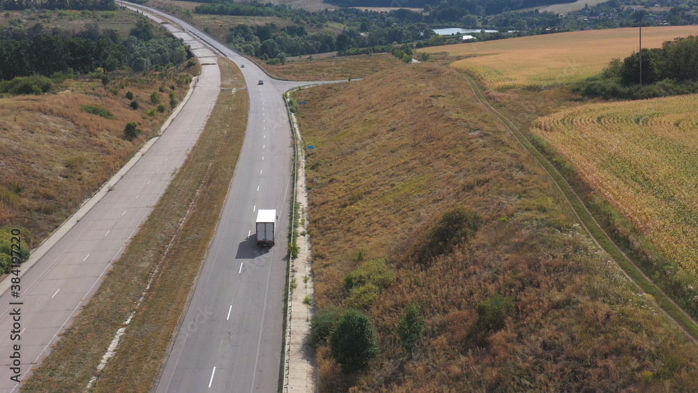 Aerial shot of truck with cargo trailer driving on empty road and transporting goods. Flying over delivery lorry moving along highway passing in beautiful countryside scene. Scenic landscape. Top view