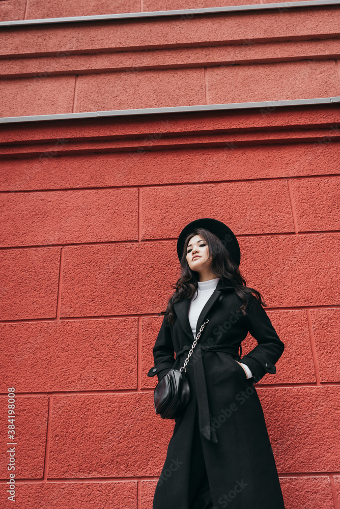 Naklejka premium Young asian girl in dark coat and hat standing in front of red wall