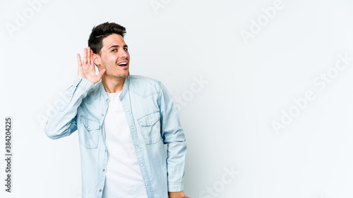 Young caucasian man isolated on white background trying to listening a gossip.