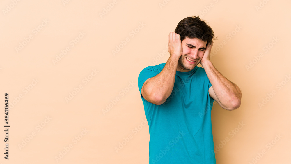 Young man isolated on beige background covering ears with hands. Young man isolated on beige background covering ears with hands.