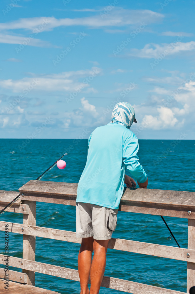 back view, medium distance of an African-American, young, male, fishing off a tropical wood pier, in blue sun protective cloths and face mask, on a sunny day, on gulf of Mexico