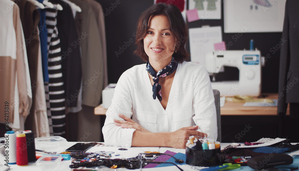 Portrait of woman seamstress sitting at table with arms folded and smiling at camera in workshop