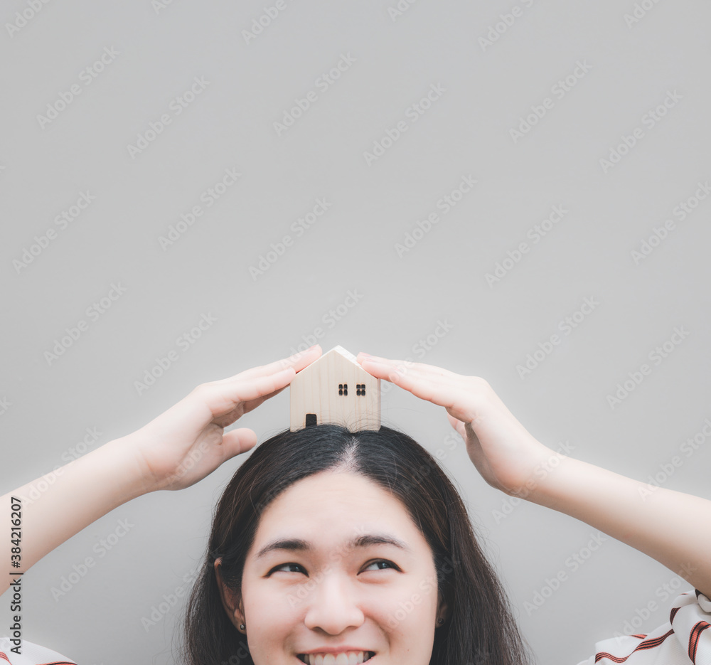 Asian woman smiling and put wooden house model on her head with two hands holding. Concept of dreaming for house loan, thinking of opportunity and home investment.